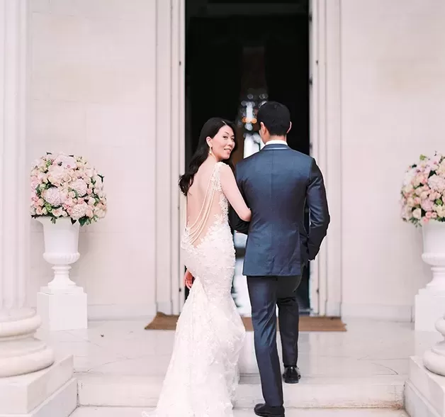Bride in backless dress and groom in suit walking arm-in-arm through entrance of white building | Quintessentially Weddings